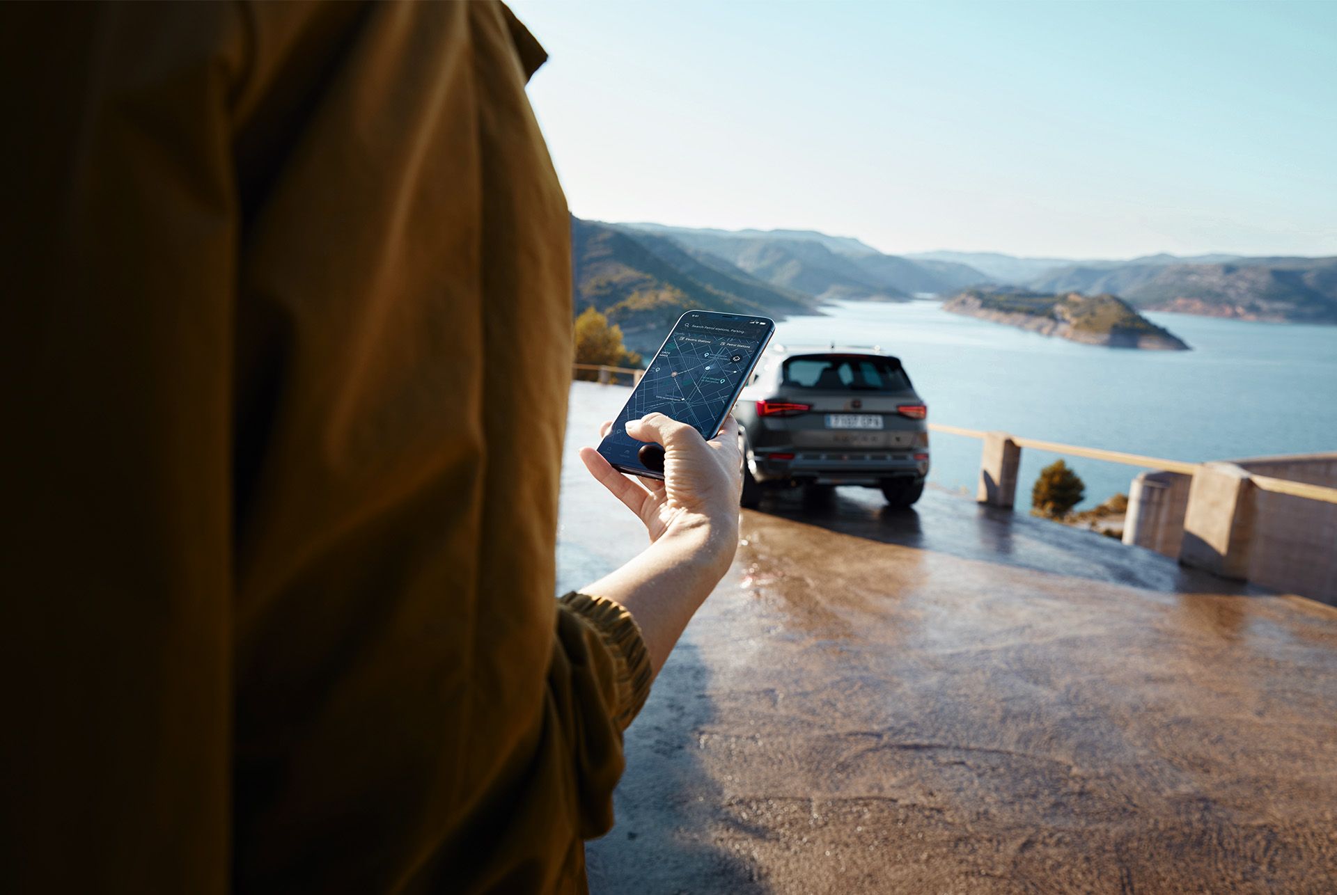 Una persona con uno smartphone e la CUPRA Ateca sullo sfondo, parcheggiata davanti a uno scenario pittoresco con vista sulle montagne e sull’acqua, come simbolo di un mondo interconnesso.