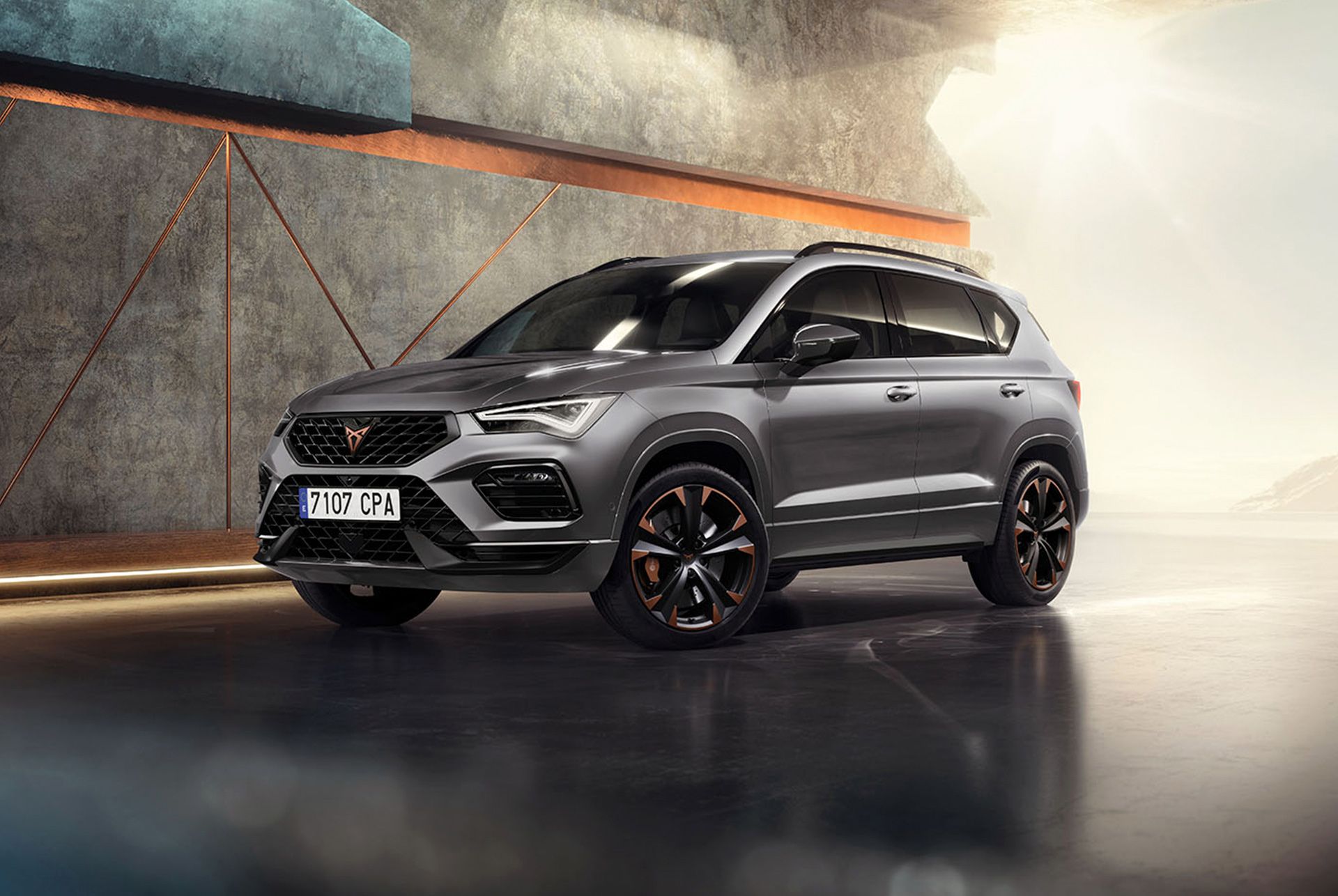 Ein CUPRA Ateca 2024 in Graphite Grey, ein sportlicher und kompakter SUV in einer modernen, industriellen Umgebung. Das Fahrzeugdesign zeichnet sich durch aerodynamische Linien, LED-Scheinwerfer und markante Leichtmetallräder mit Kupferakzenten aus. Der Hintergrund zeigt ein geometrisches Wanddesign, das im Kontrast zum eleganten Fahrzeugdesign steht.